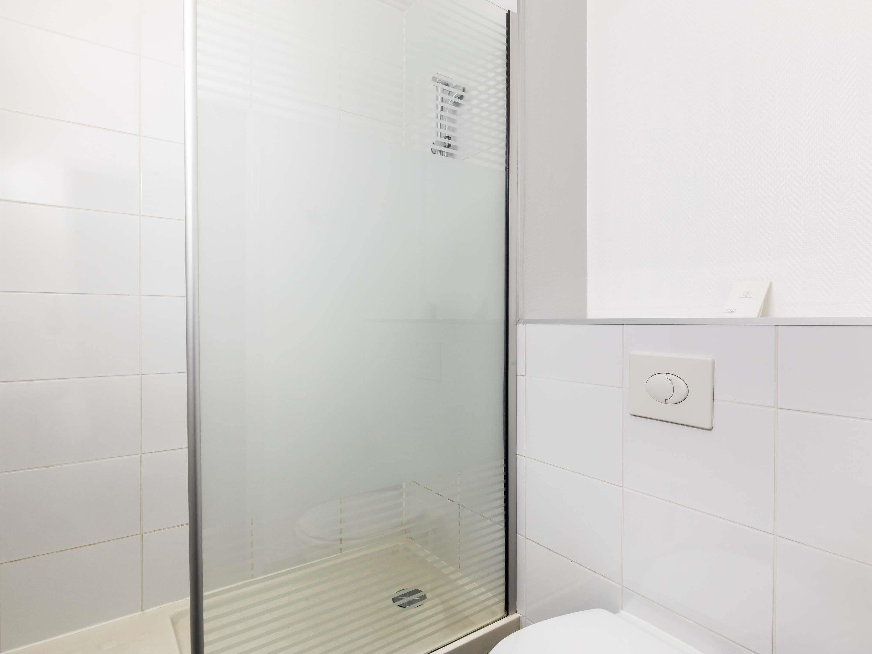 Un baño moderno con cabina de ducha de vidrio, paredes con azulejos blancos e interruptor de luz a la vista.