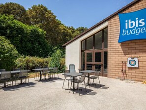 Un espacio al aire libre con mesas y sillas, un edificio con un letrero azul y un dardo en la pared.