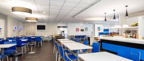 Una cafetería moderna con sillas azules y mesas blancas, una barra con productos alimenticios y un televisor de pantalla plana en la pared.