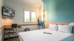 Quarto de hotel com cama, escrivaninha com laptop, cadeira, televisão fixada na parede e janela.