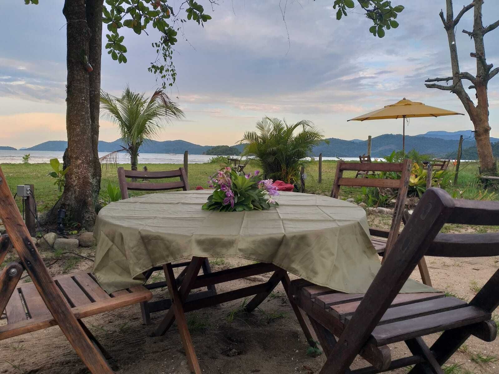 A round table with a tablecloth, surrounded by wooden chairs, set outdoors with a view of a body of water and mountains in the background.