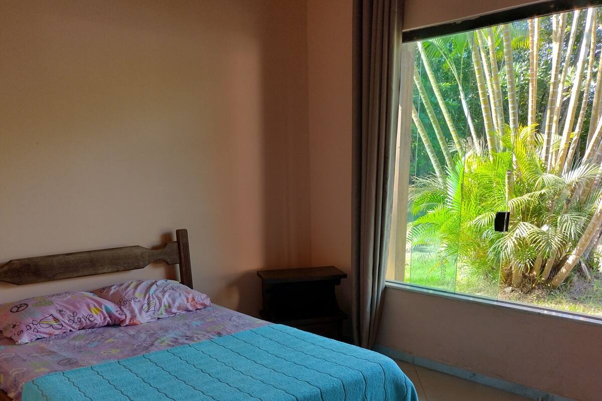 A bedroom with a bed, a nightstand, a window with a view of greenery, and a chair.