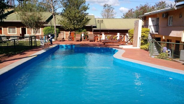 A swimming pool with a wooden deck and lounge chairs, surrounded by buildings with a green roof. - Nuevo Raices Apart (Puerto Iguazú)