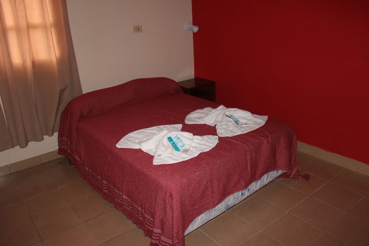 A single bed with a red bedspread and white towels, a nightstand, and a red wall.