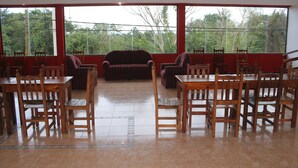 A dining area with wooden tables and chairs, large windows with a view of trees, and a red wall. - Nuevo Raices Apart (Puerto Iguazú)