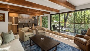 Ein modernes Wohnzimmer mit Sofa, Sesseln, einem Couchtisch und Blick auf eine Küche sowie eine Sitzgelegenheit im Freien durch große Fenster.