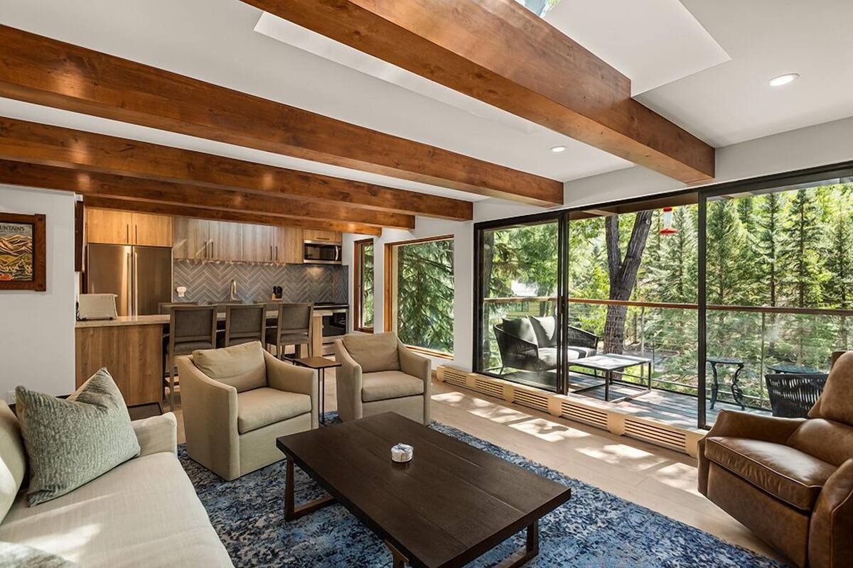 Ein modernes Wohnzimmer mit Sofa, Sesseln, einem Couchtisch und Blick auf eine Küche sowie eine Sitzgelegenheit im Freien durch große Fenster.