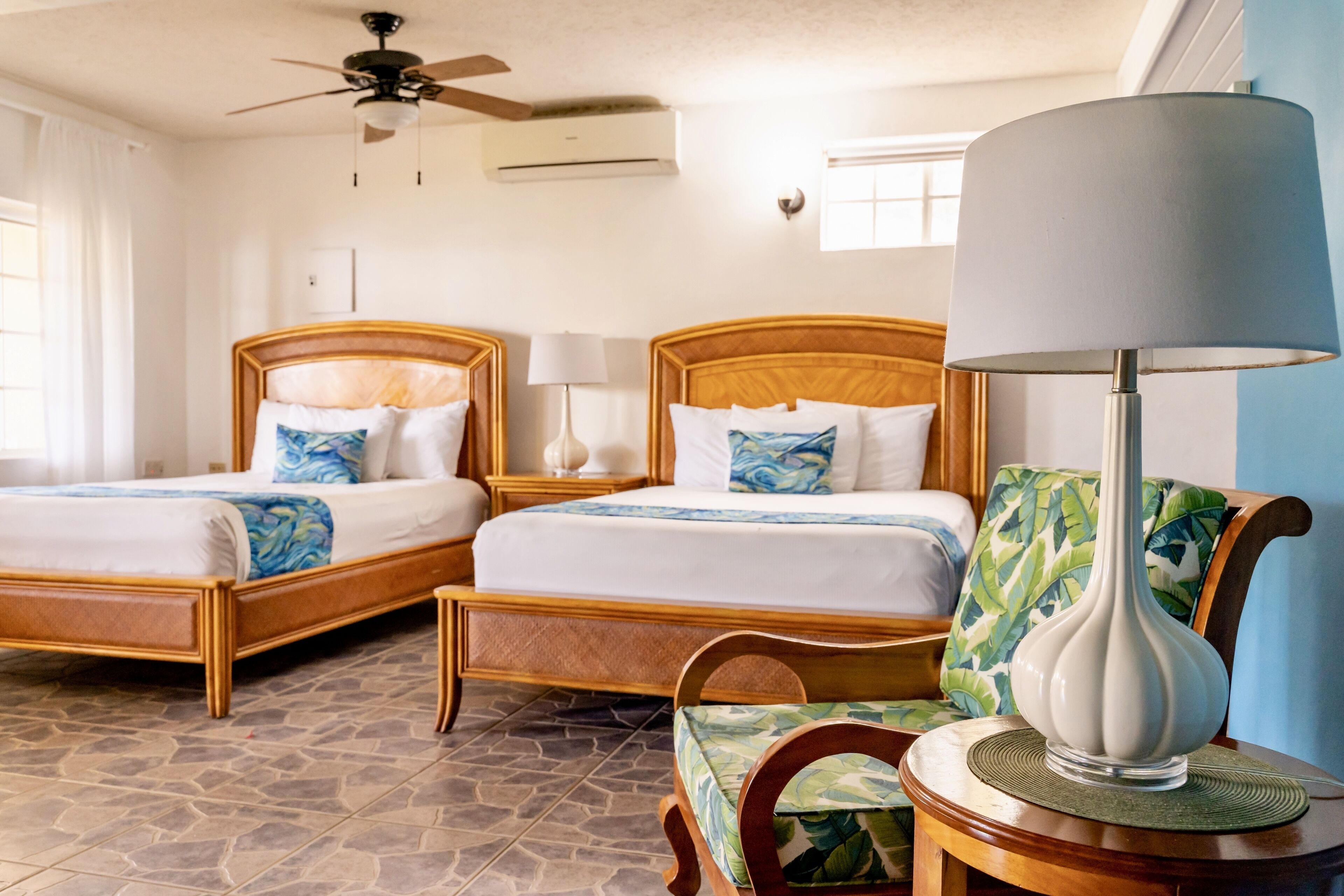 Une chambre d’hôtel avec deux lits en bois, un ventilateur de plafond, un appareil de climatisation, une lampe et une chaise avec un coussin à motifs de feuilles.