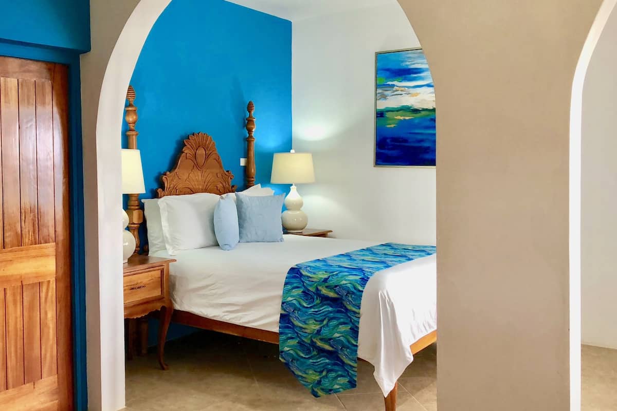 A bedroom with a bed, a wooden headboard, two bedside tables with lamps, a blue wall, and a painting.