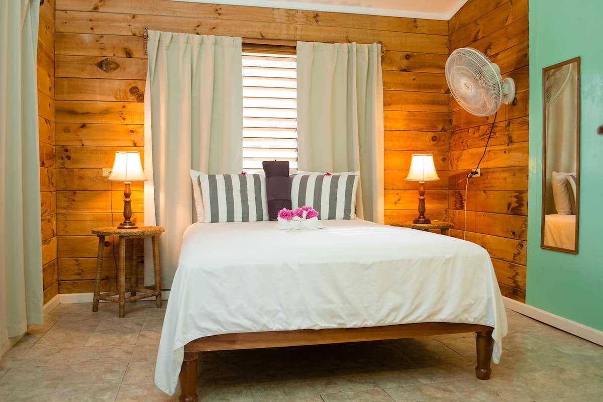 A wooden-paneled bedroom with a bed, two bedside lamps, a fan, and a window with curtains.