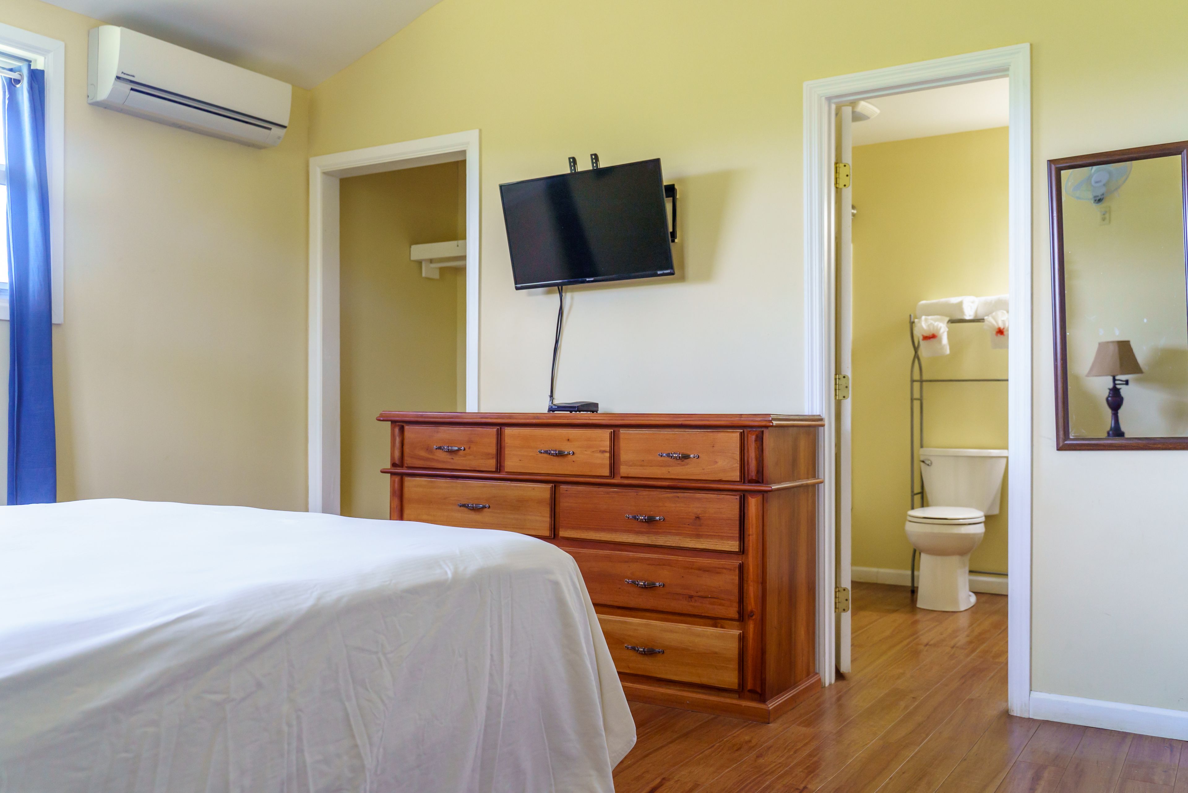 Une chambre à coucher avec un lit, une commode en bois, un téléviseur fixé au mur et une salle de bain visible à travers une porte ouverte.
