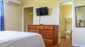 Une chambre à coucher avec un lit, une commode en bois, un téléviseur fixé au mur et une salle de bain visible à travers une porte ouverte.