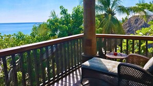 A wooden deck with a wicker sofa and a small round table, overlooking a tropical landscape with palm trees and a view of the ocean. - The Westender Inn (Negril)
