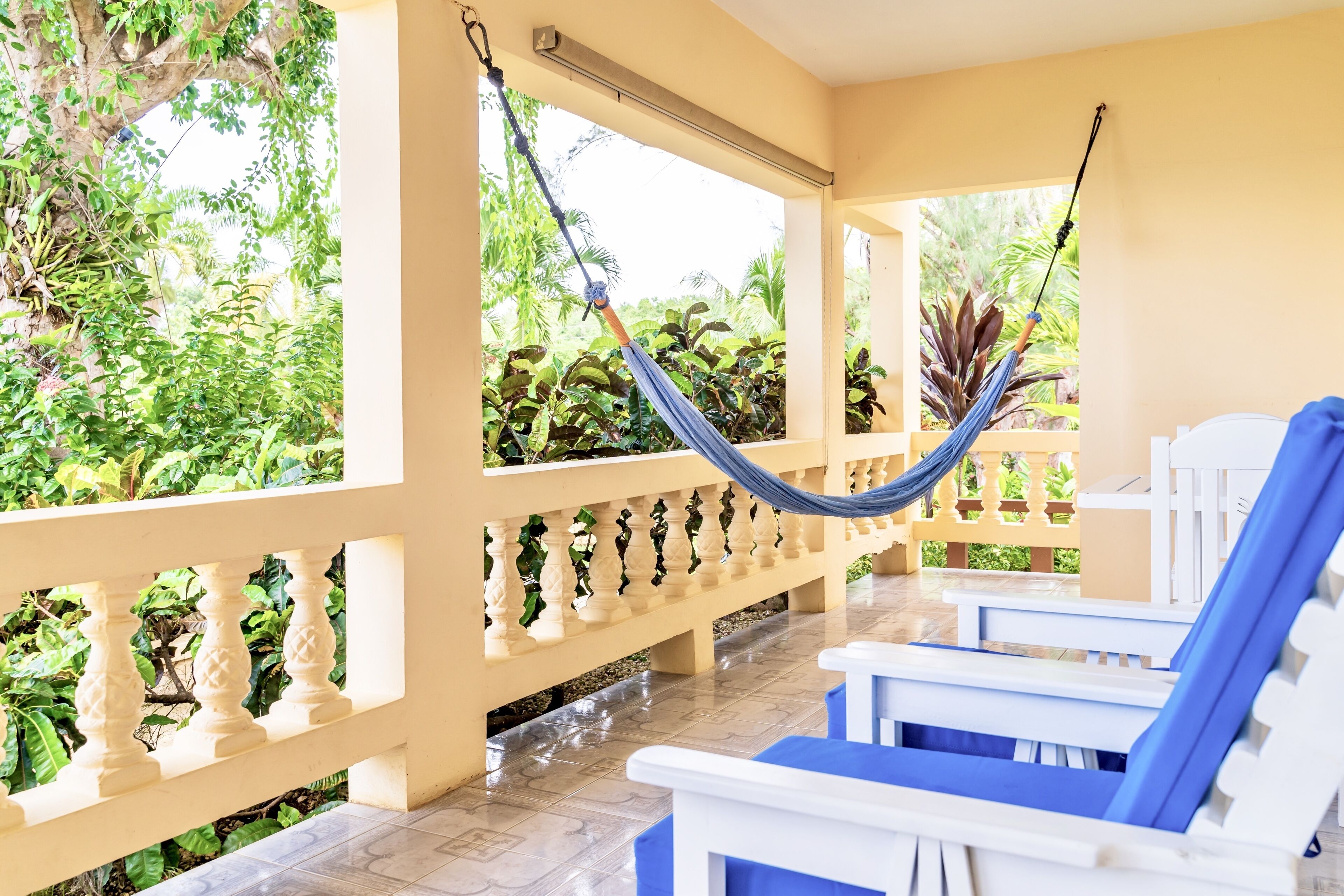 A covered outdoor seating area with white and blue lounge chairs, a hammock, and a view of lush greenery.
