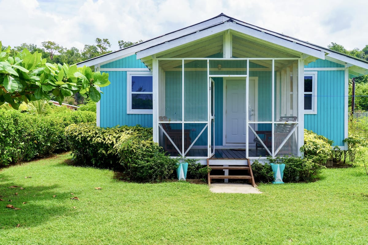 A small, single-story house with a porch, surrounded by lush greenery.
