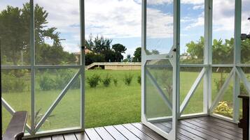 A covered outdoor area with a wooden deck, a view of a grassy field, and a building in the background.