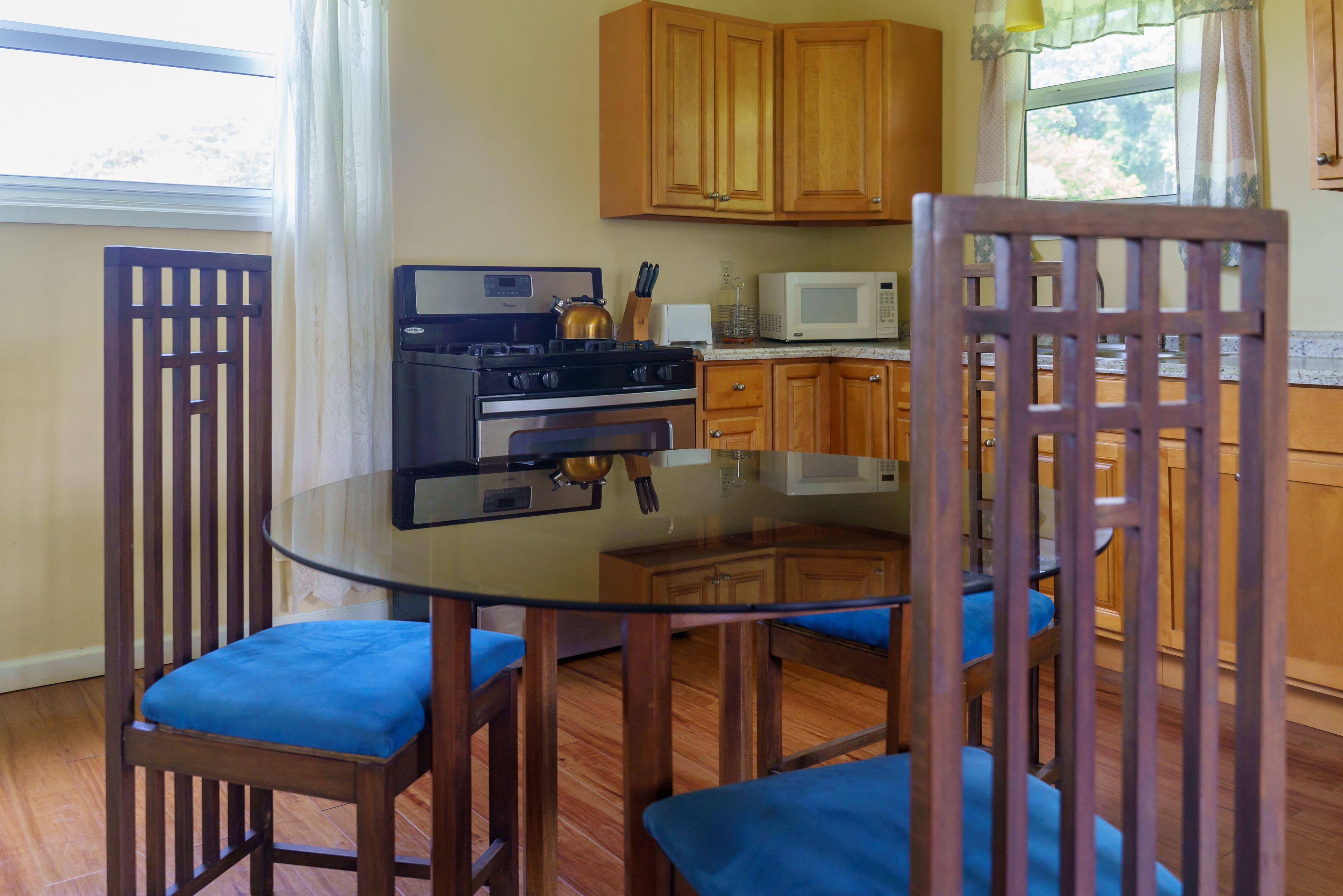 Una cocina con armarios de madera, una estufa a gas, un microondas y una mesa de comedor de vidrio con sillas azules.