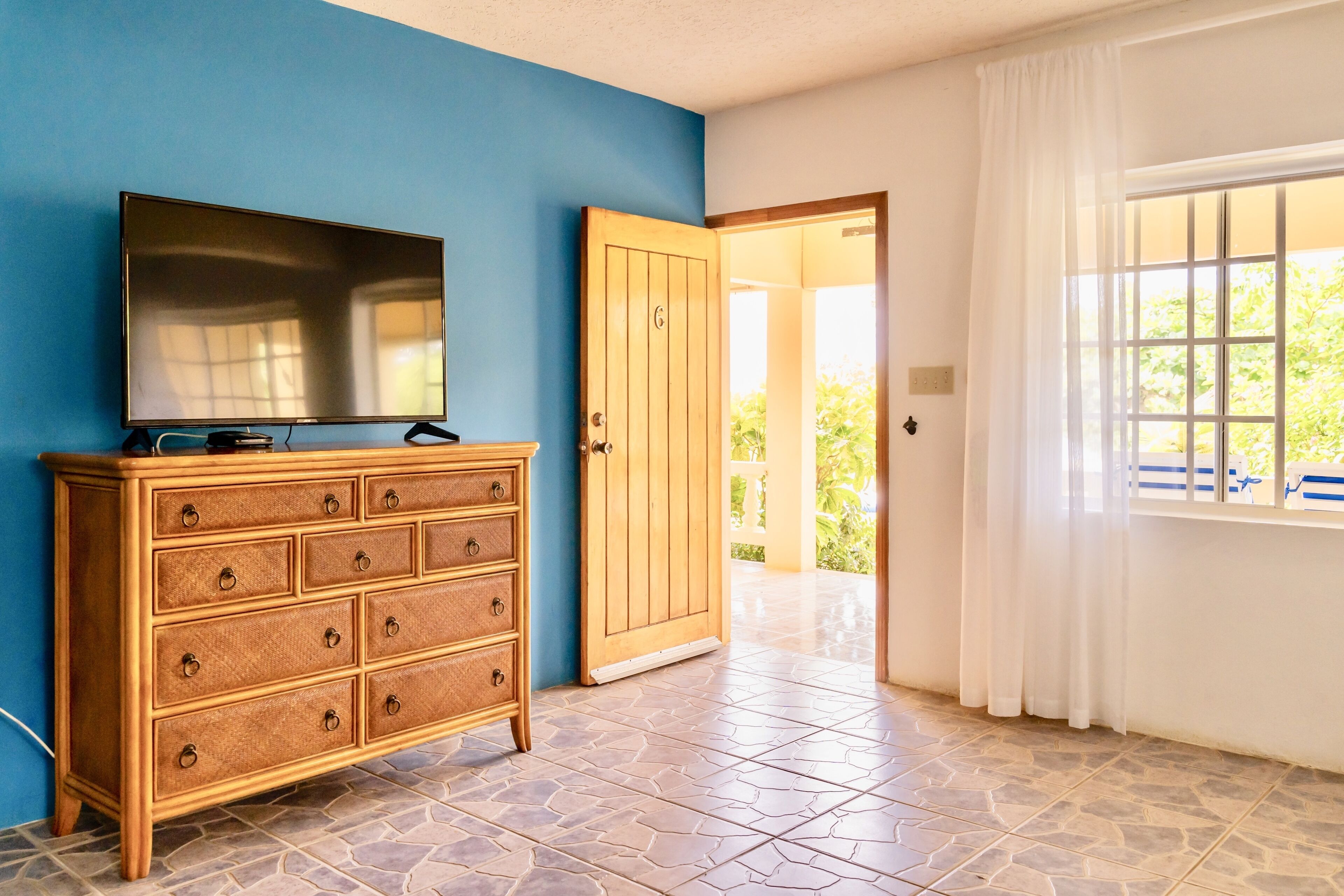 A room with a wooden dresser, a television on top, a tiled floor, a door leading outside, and a window with curtains.