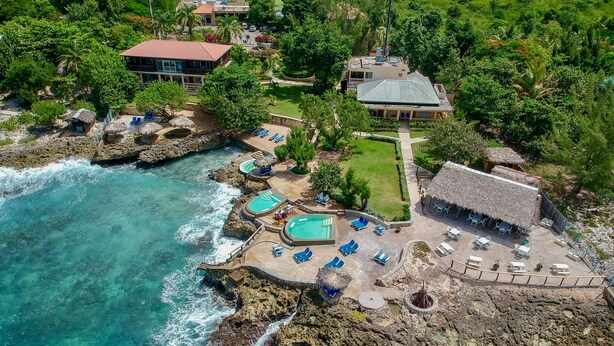 An aerial view of a coastal resort with multiple pools, lounge areas, and a thatched-roof building.