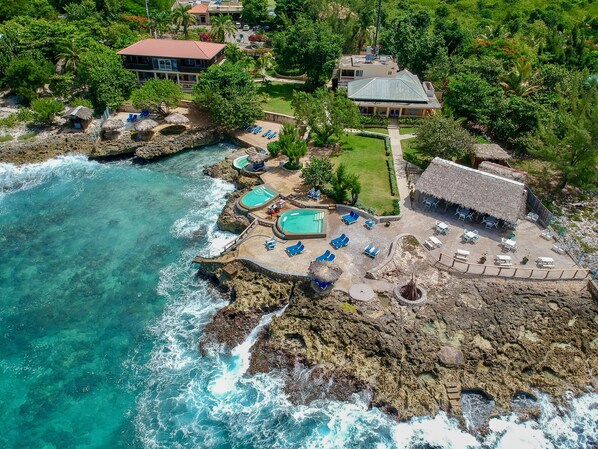 An aerial view of a coastal resort with multiple pools, lounge areas, and a thatched-roof building.