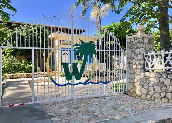 A gated entrance with a decorative emblem, a stone wall, and a paved pathway leading to a building. - The Westender Inn (Negril)