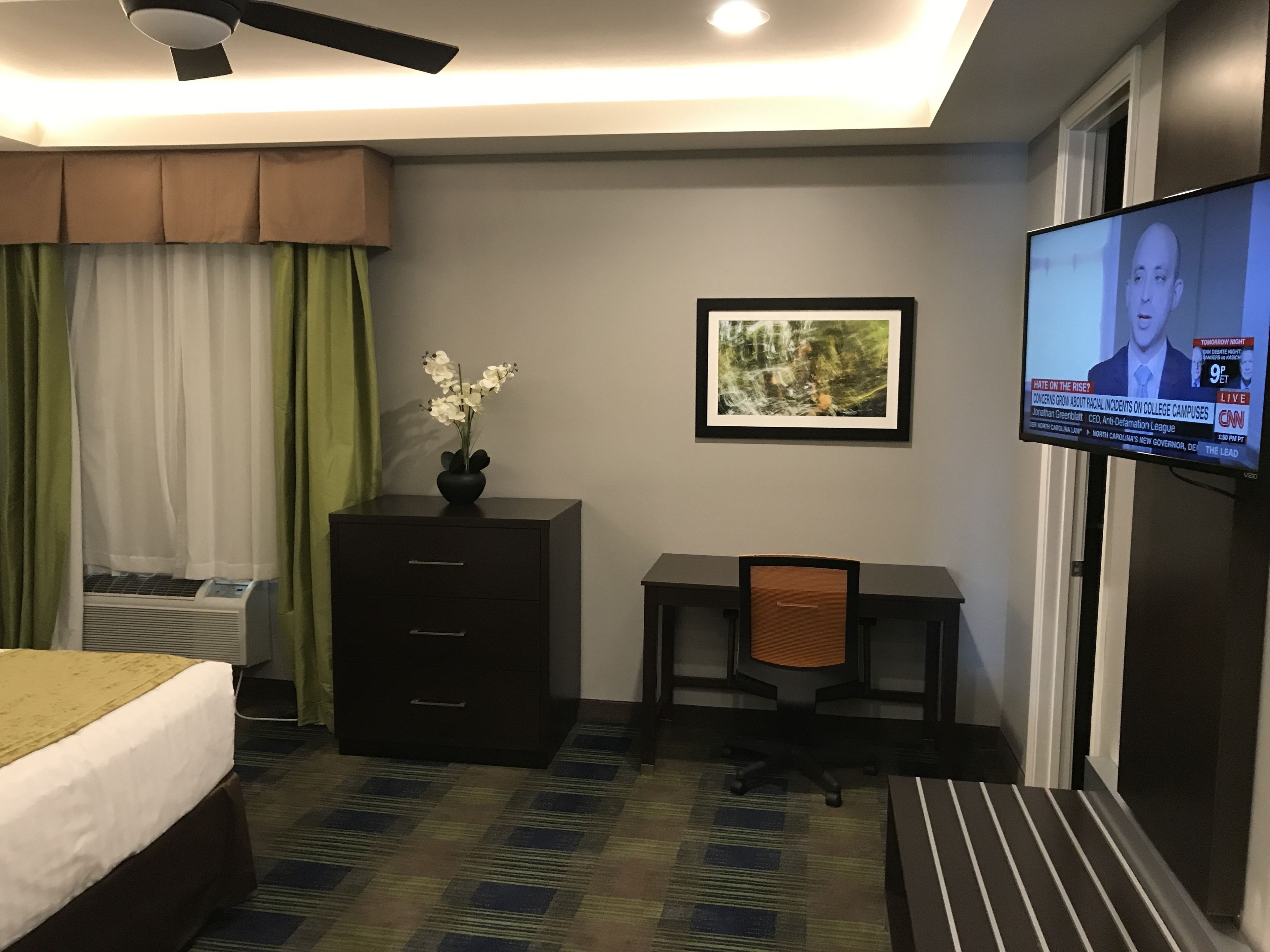 Hotelkamer met een bed, bureau, stoel, televisie, dressoir en raam met gordijnen.