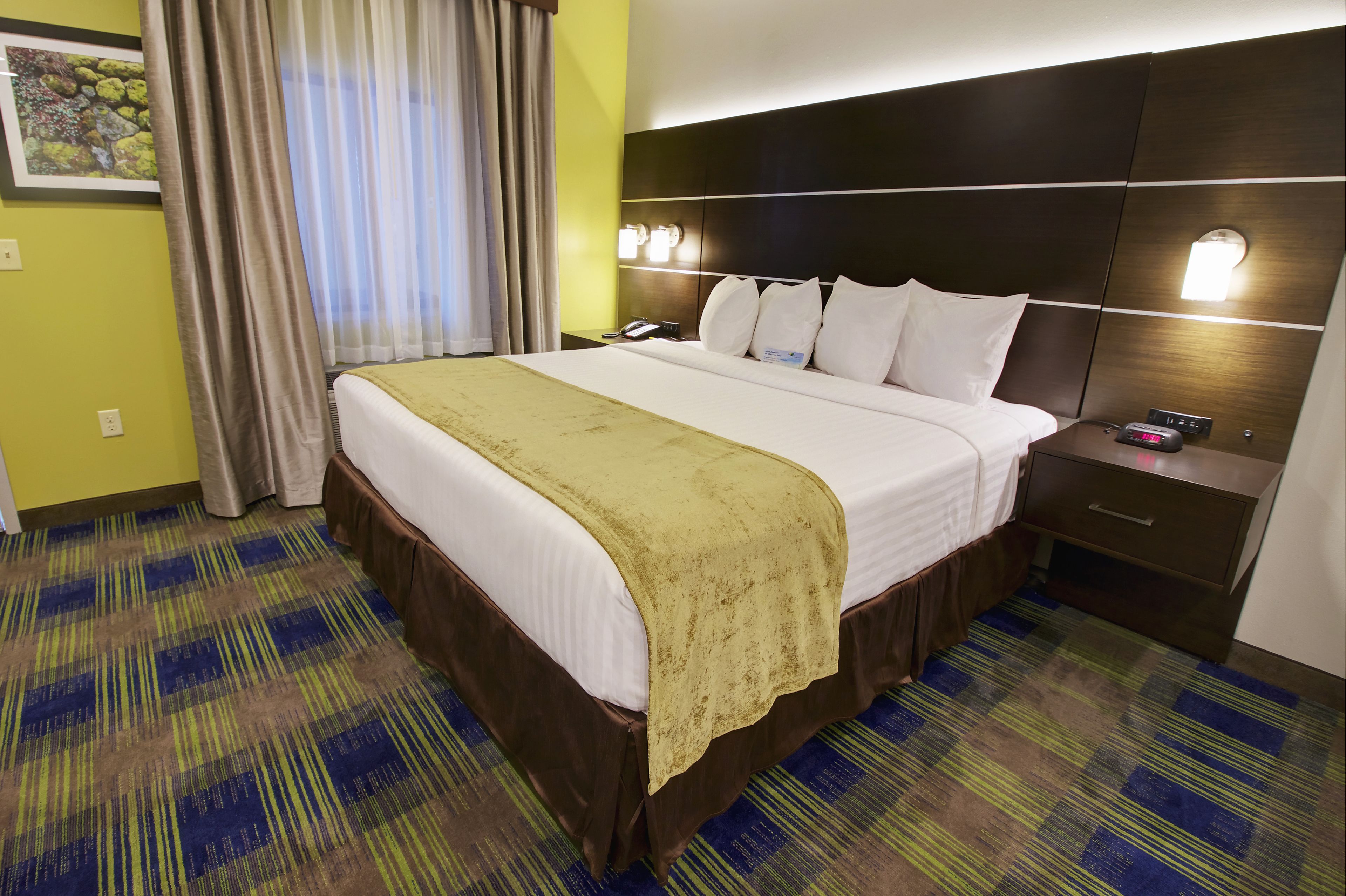 A hotel room with a large bed, a bedside table with a phone, and a nightstand with a clock.
