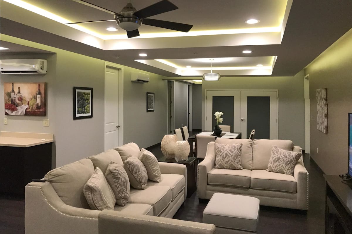 A modern living room with two beige sofas, a ceiling fan, recessed lighting, and a small table with decorative items.