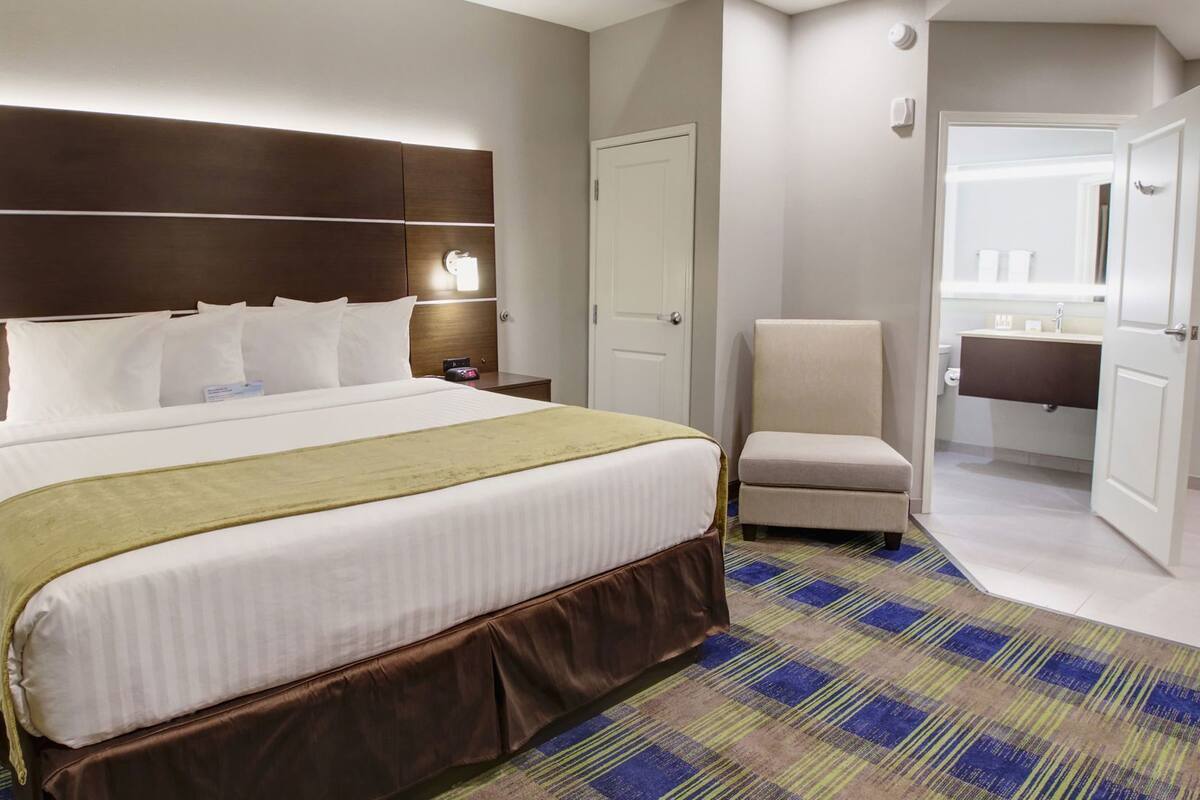 A hotel room with a large bed, a chair, a nightstand, a bedside table, and a bathroom visible through an open door.