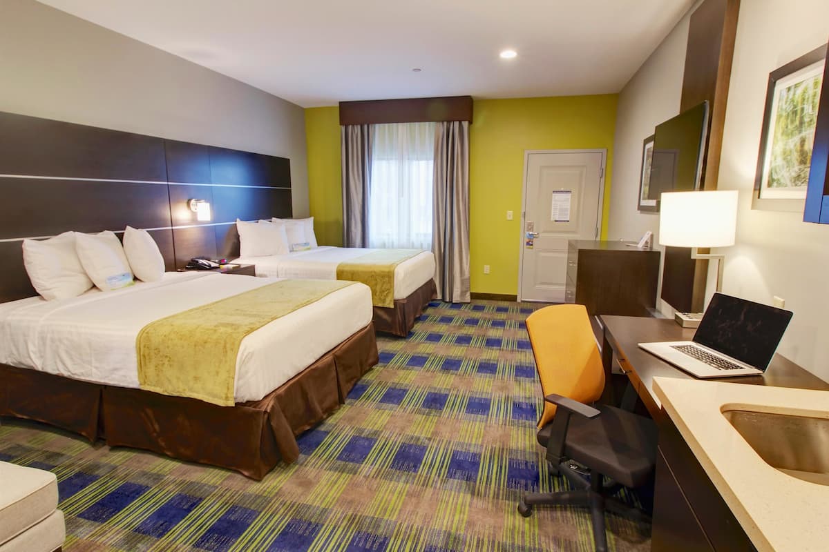 A hotel room with two beds, a desk with a laptop, a chair, and a sink.