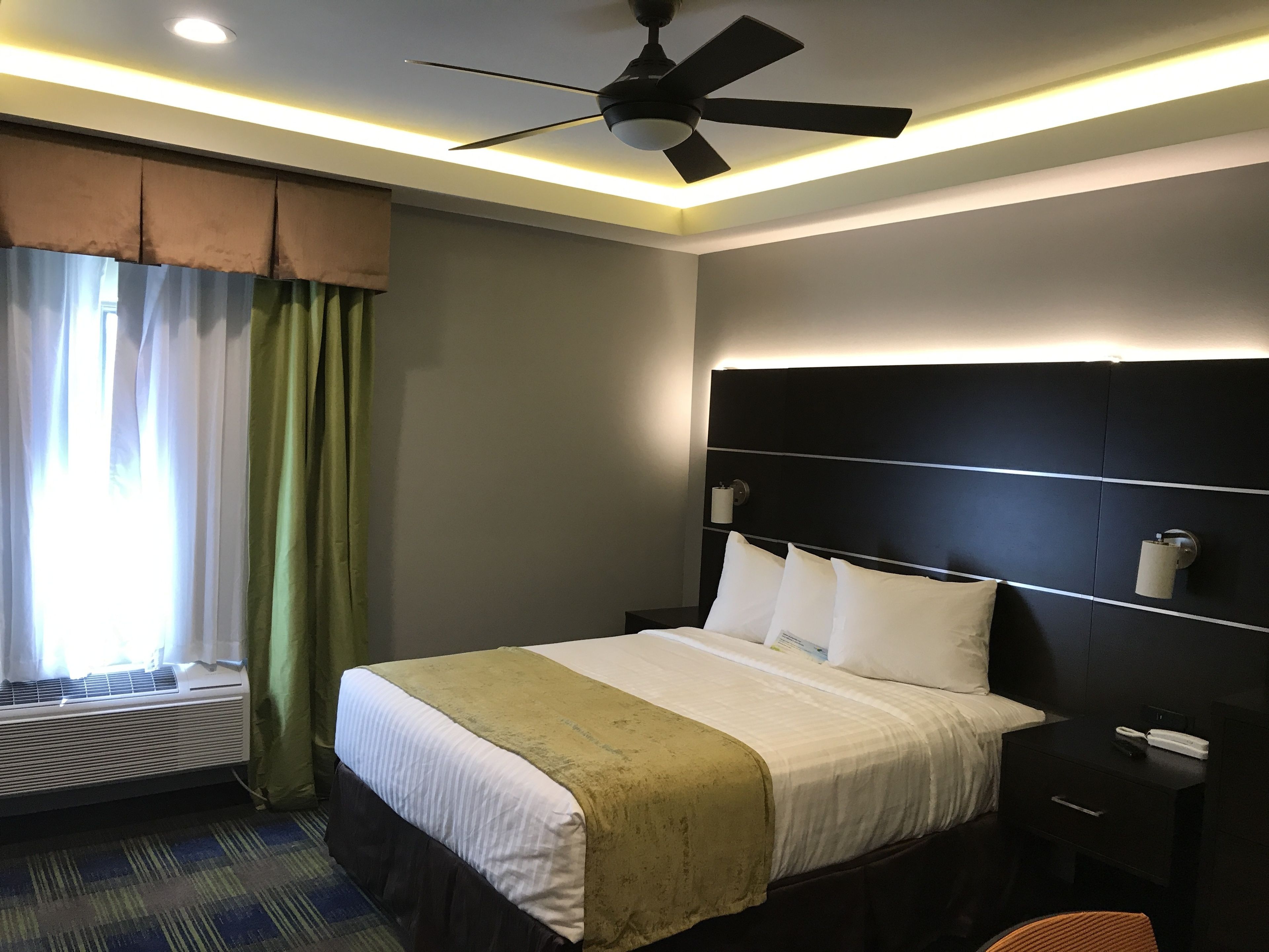 A hotel room with a bed, a nightstand, a ceiling fan, a window with curtains, and an air conditioning unit.