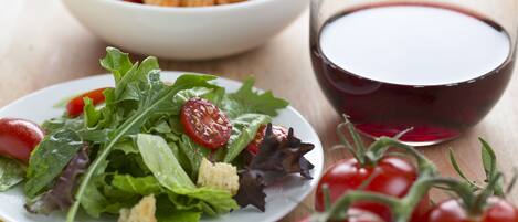 Un plato de ensalada de hojas verdes mezcladas con tomates cherry y croutons, un bol de pasta al horno y una copa de vino tinto sobre una mesa de madera.