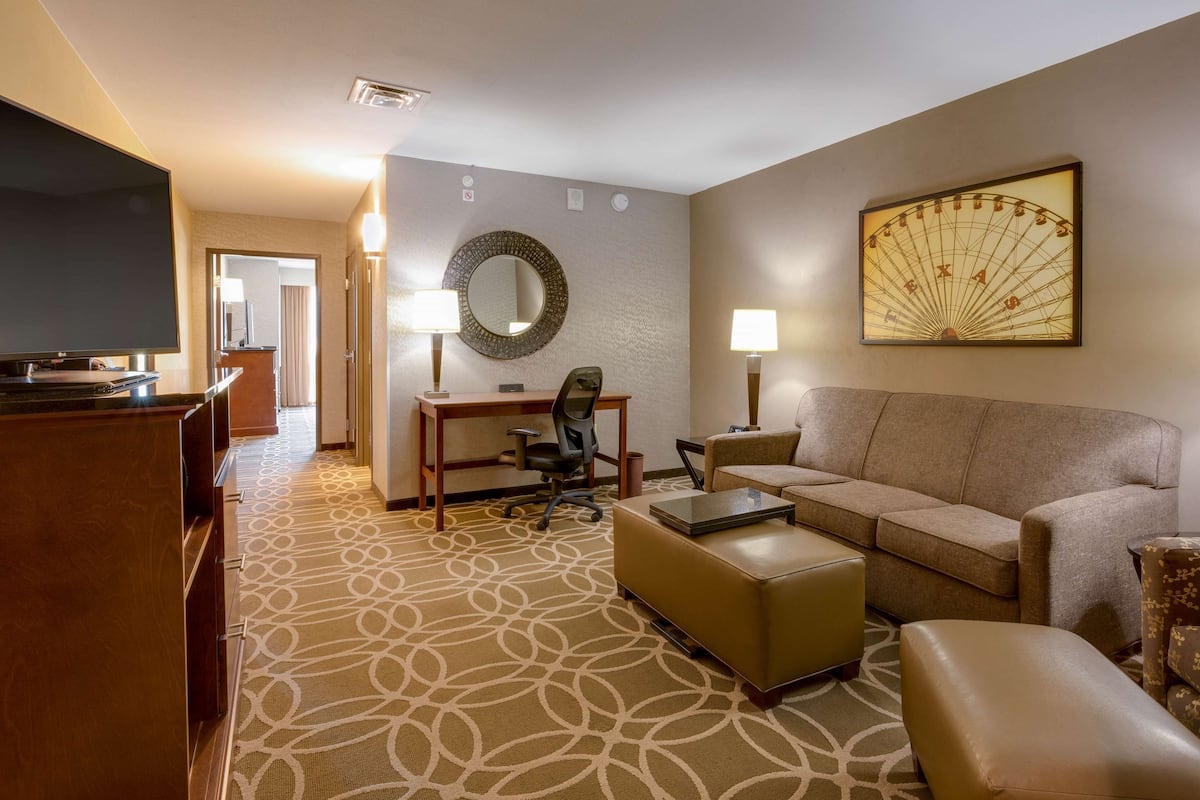 A hotel room with a large flat-screen TV, a desk with a chair, a sofa, a coffee table, and a large wall-mounted mirror.
