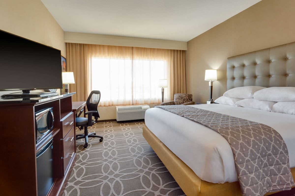 A hotel room with a large bed, a desk with a flat-screen TV, a chair, a lamp, and a window with curtains.