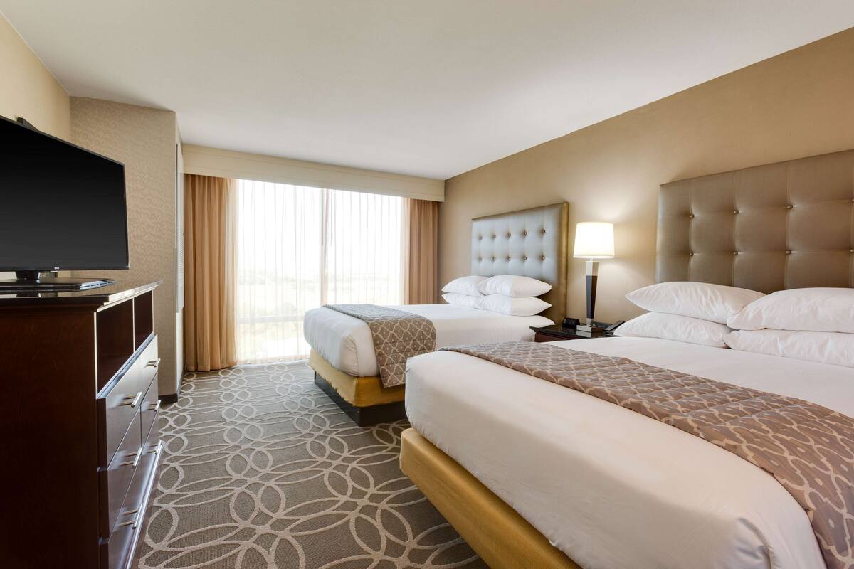 A hotel room with two beds, a TV, a desk, and a large window with curtains.