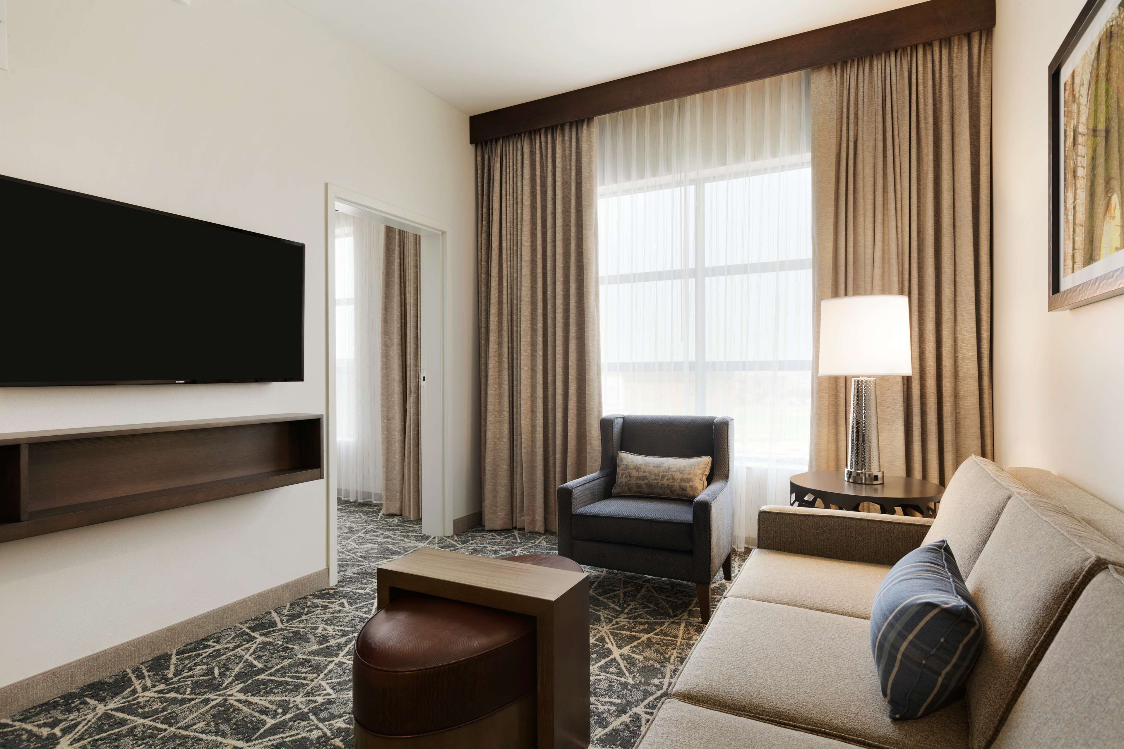 A hotel room with a flat-screen TV mounted on the wall, a sofa, a chair, a small table, and a lamp.