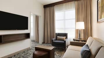 A hotel room with a flat-screen TV mounted on the wall, a sofa, a chair, a small table, and a lamp.