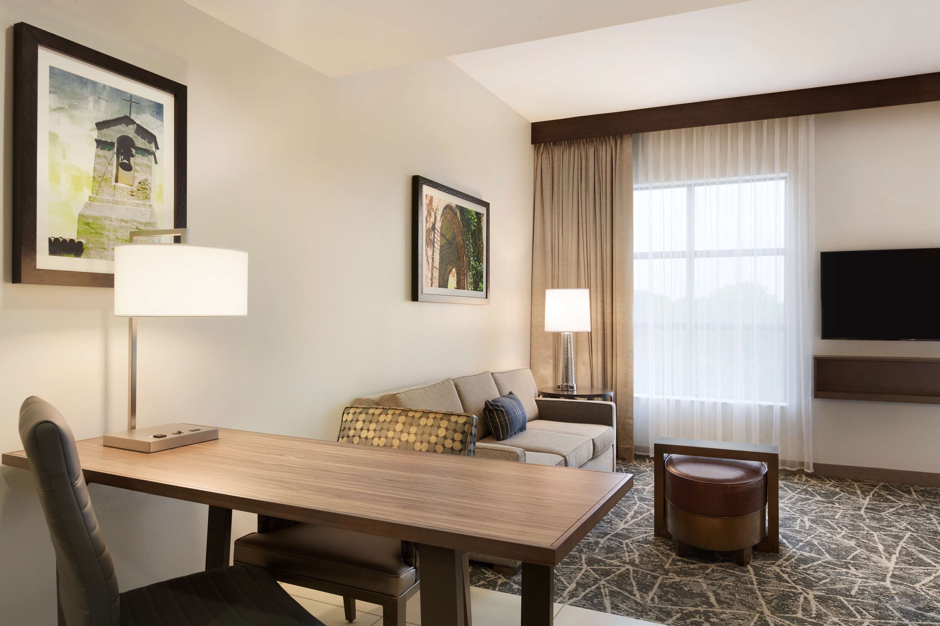 A hotel room with a wooden dining table, a sofa, a TV mounted on the wall, and a large window with curtains.