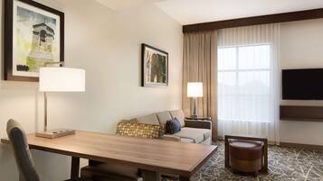 A hotel room with a wooden dining table, a sofa, a TV mounted on the wall, and a large window with curtains.