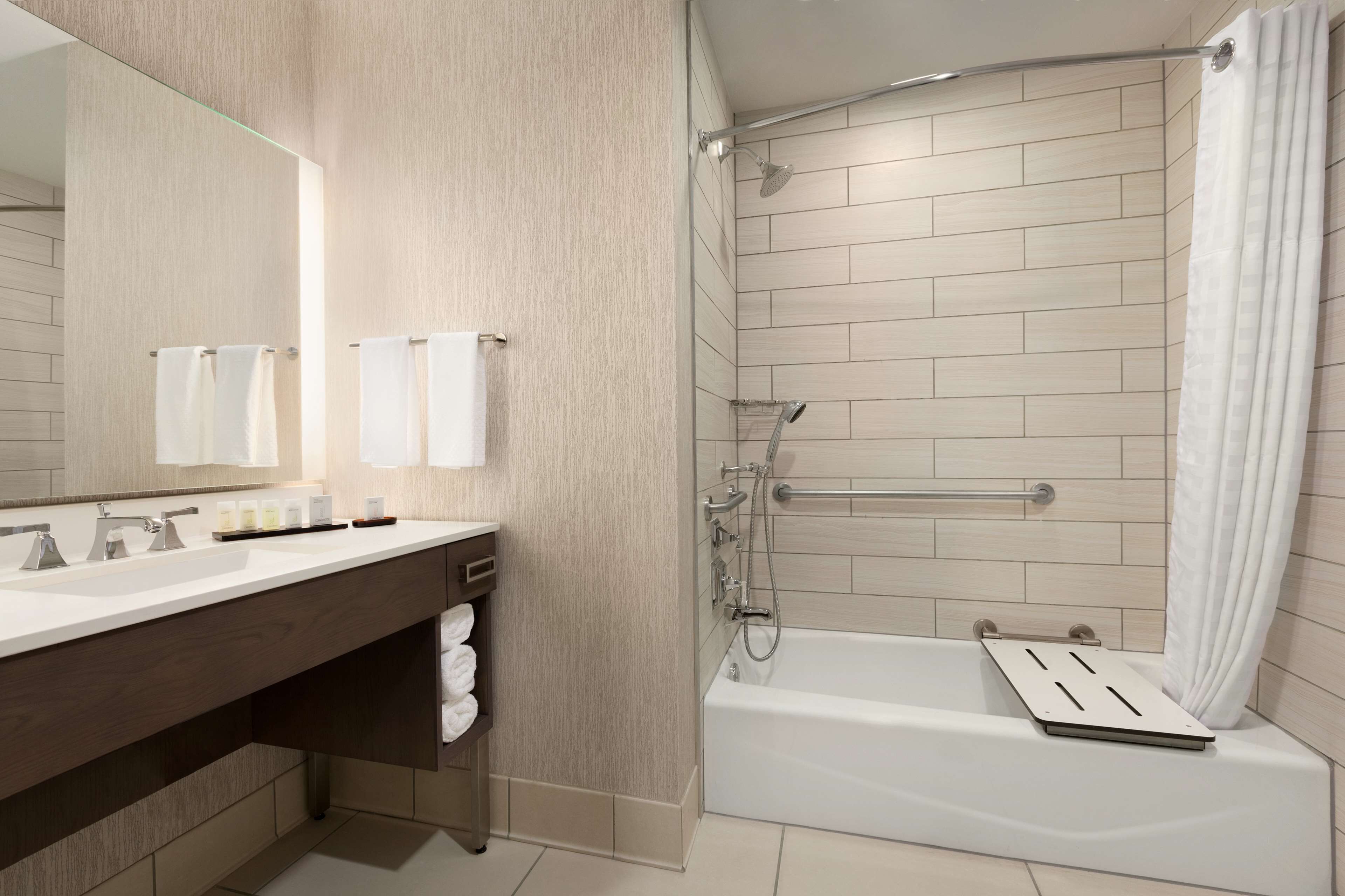 A modern bathroom with a dual sink vanity, a large mirror, a shower with a handheld showerhead, and a bathtub with a shower seat.