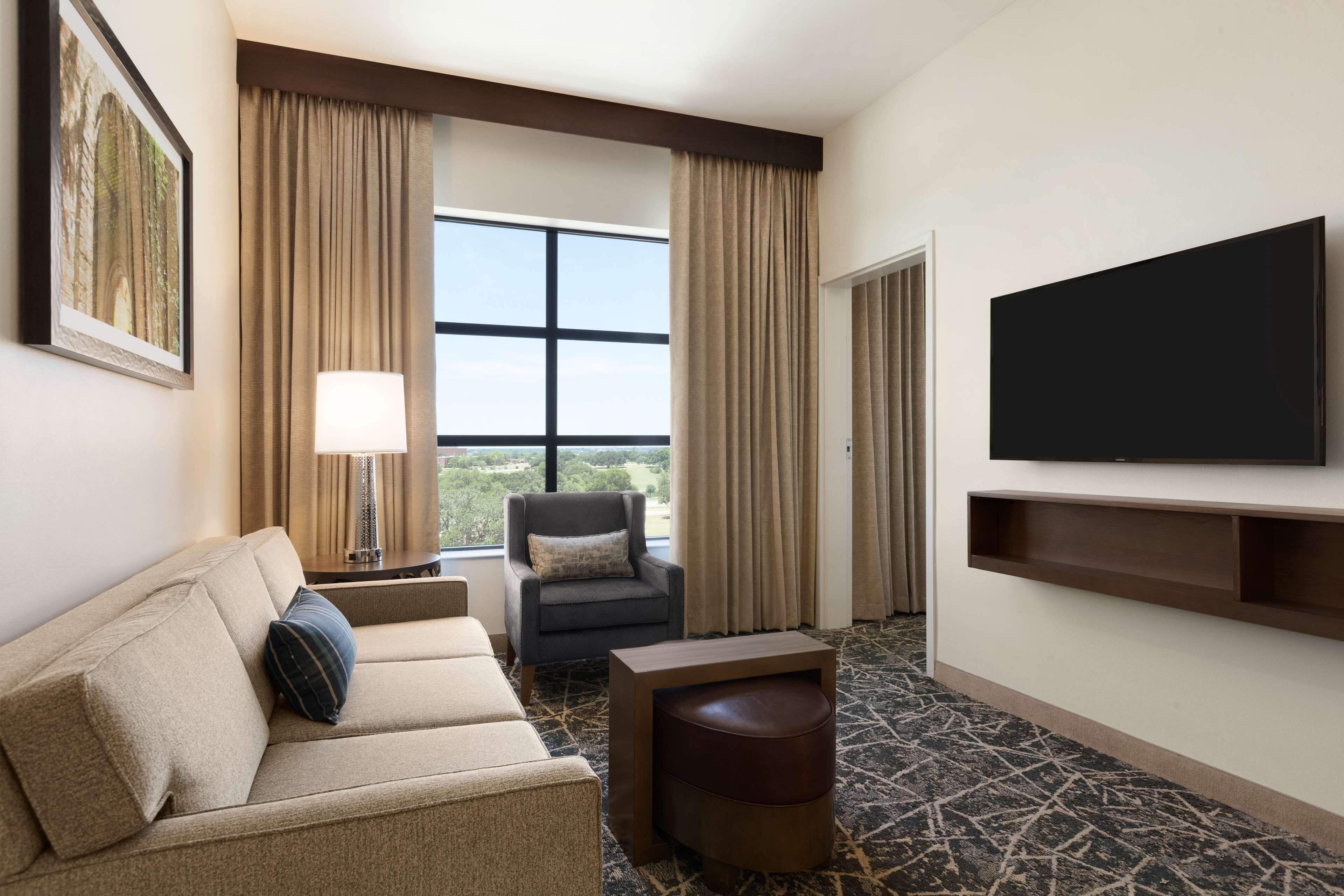 A hotel room with a sofa, armchair, TV, and a large window with curtains.