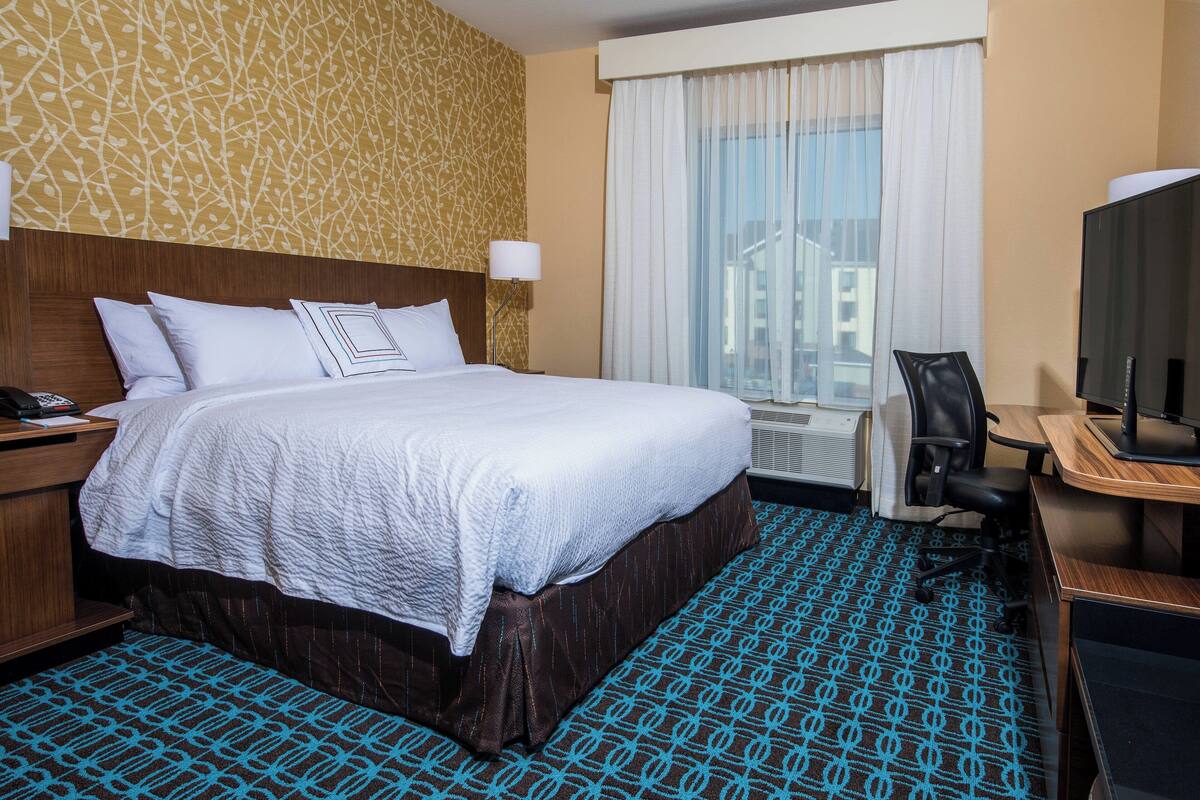 Habitación de hotel con una cama grande, un escritorio con silla, televisión y ventana con cortinas.