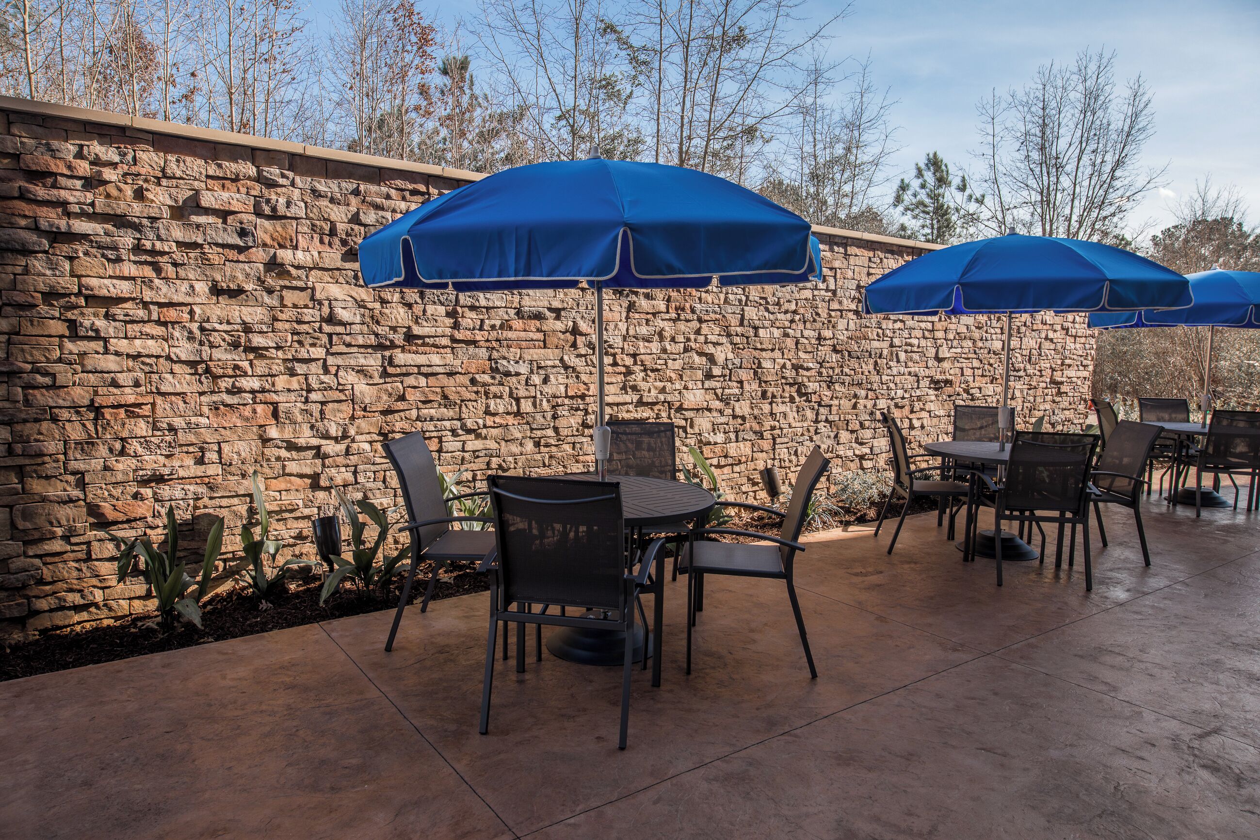 An outdoor patio area with stone walls, metal chairs, and blue umbrellas.