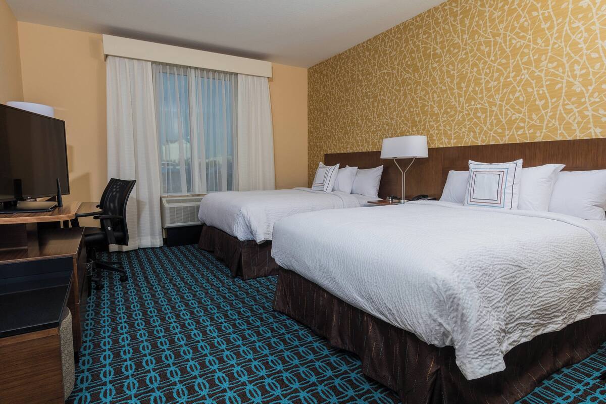 Habitación de hotel con dos camas, un escritorio con silla, televisor y alfombra estampada.