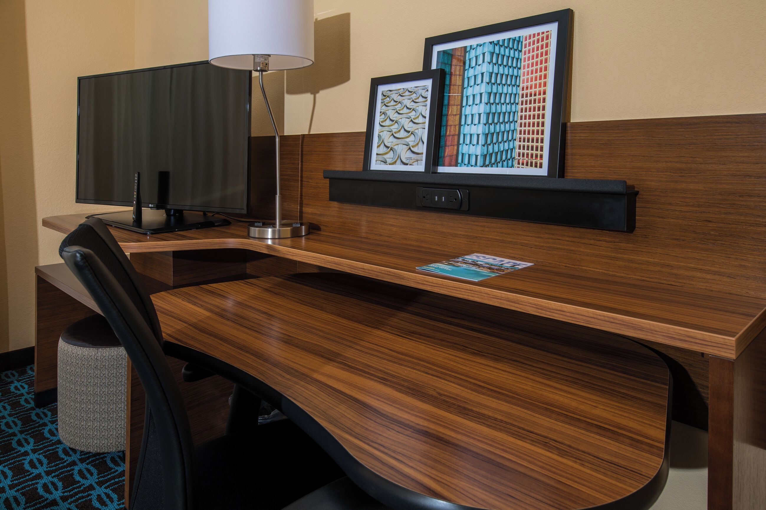 A hotel room with a wooden desk, a flat-screen TV, a lamp, and two framed pictures on the wall.