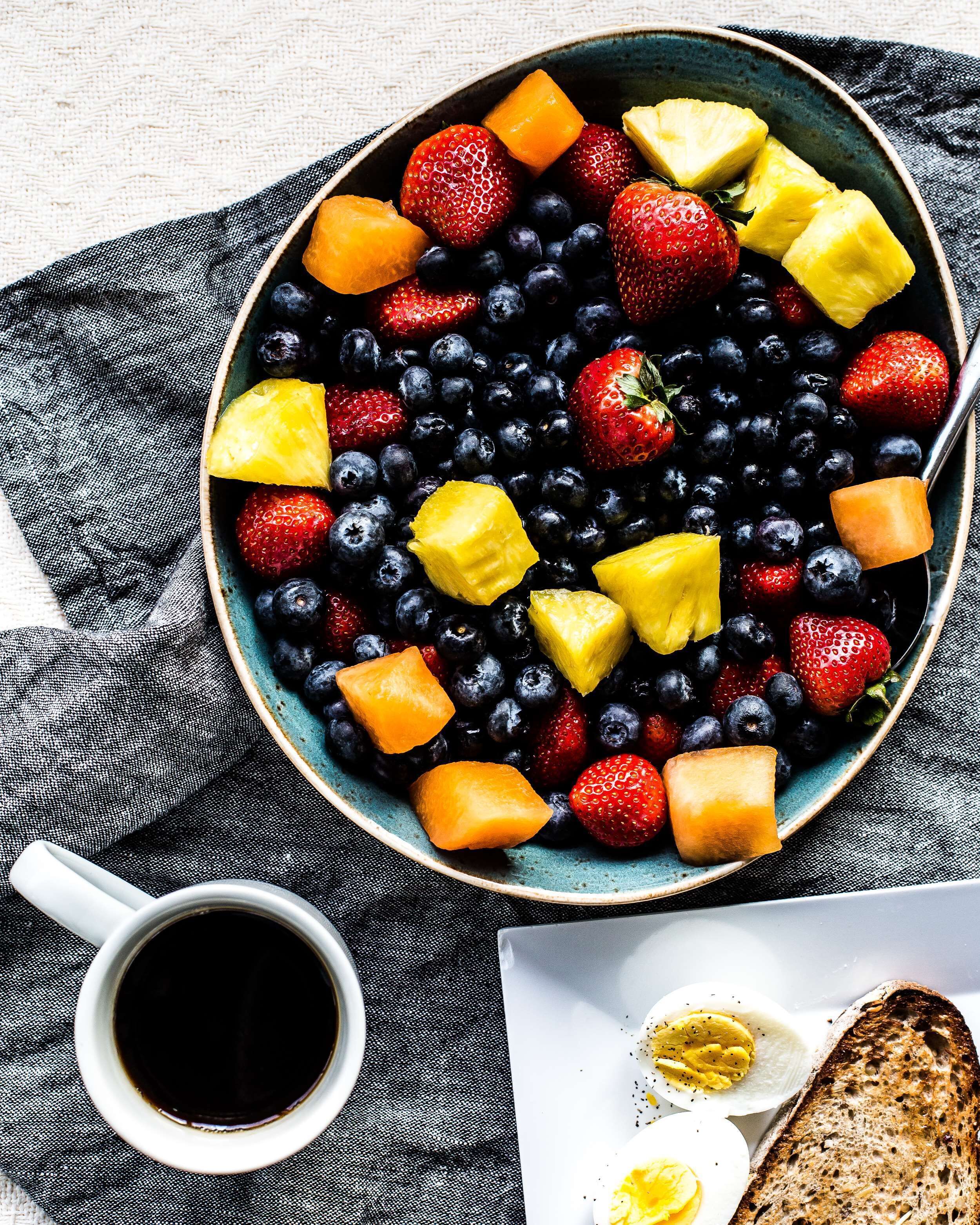 A bowl of mixed fruits including strawberries, blueberries, and pineapple chunks, accompanied by a cup of coffee and toast.