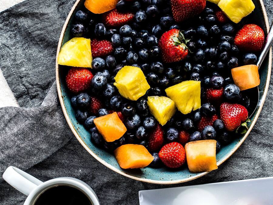 A bowl of mixed fruits including strawberries, blueberries, and pineapple chunks, accompanied by a cup of coffee and toast.