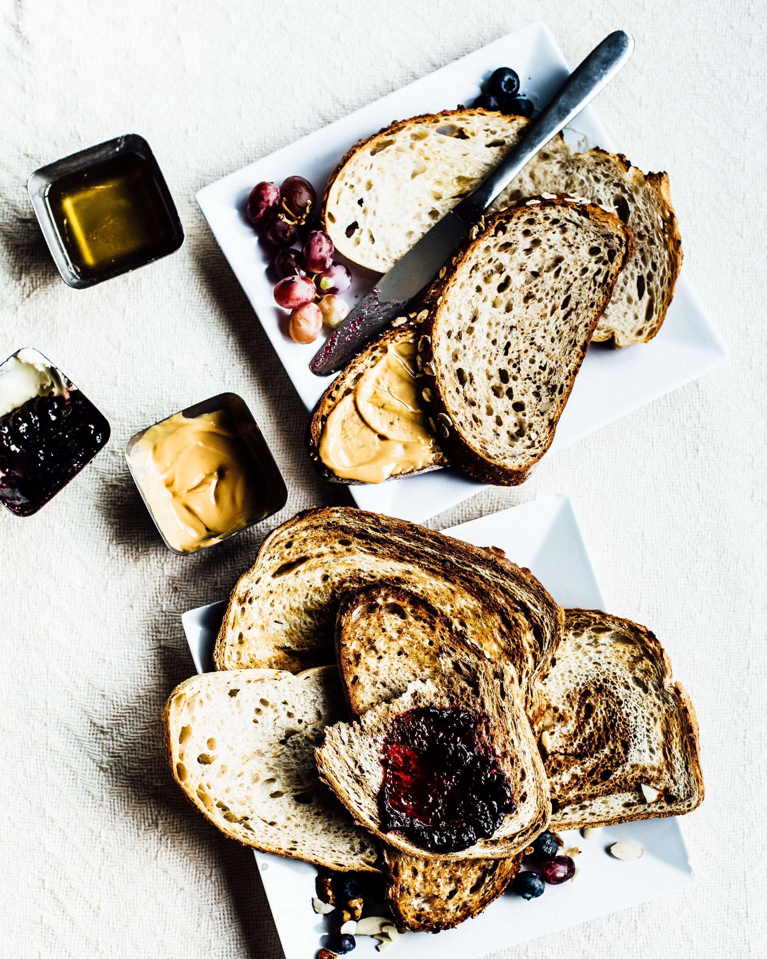 Slices of whole grain bread with peanut butter and jam, accompanied by grapes and a knife on a white plate.