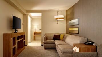 A modern hotel room with a sectional sofa, a flat-screen TV on a wooden console, a bedside table with a lamp, and a framed picture on the wall.