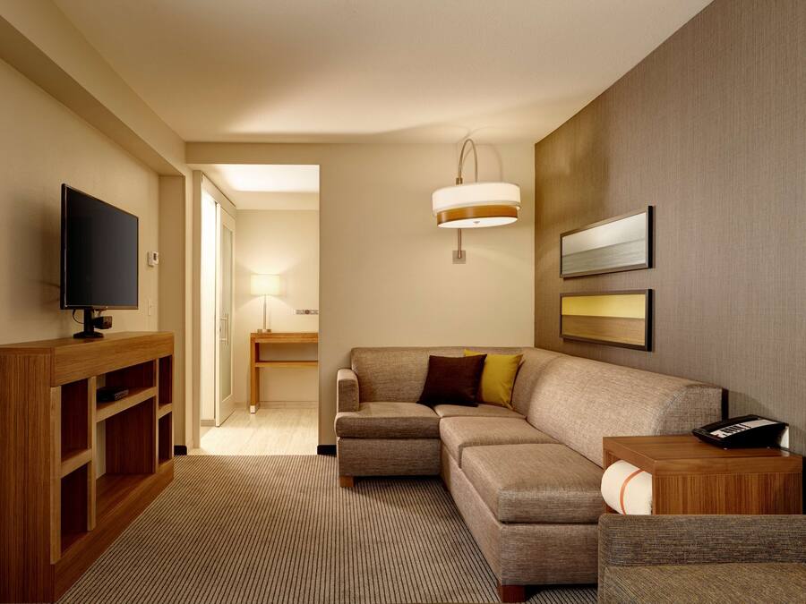 A modern hotel room with a sectional sofa, a flat-screen TV on a wooden console, a bedside table with a lamp, and a framed picture on the wall.